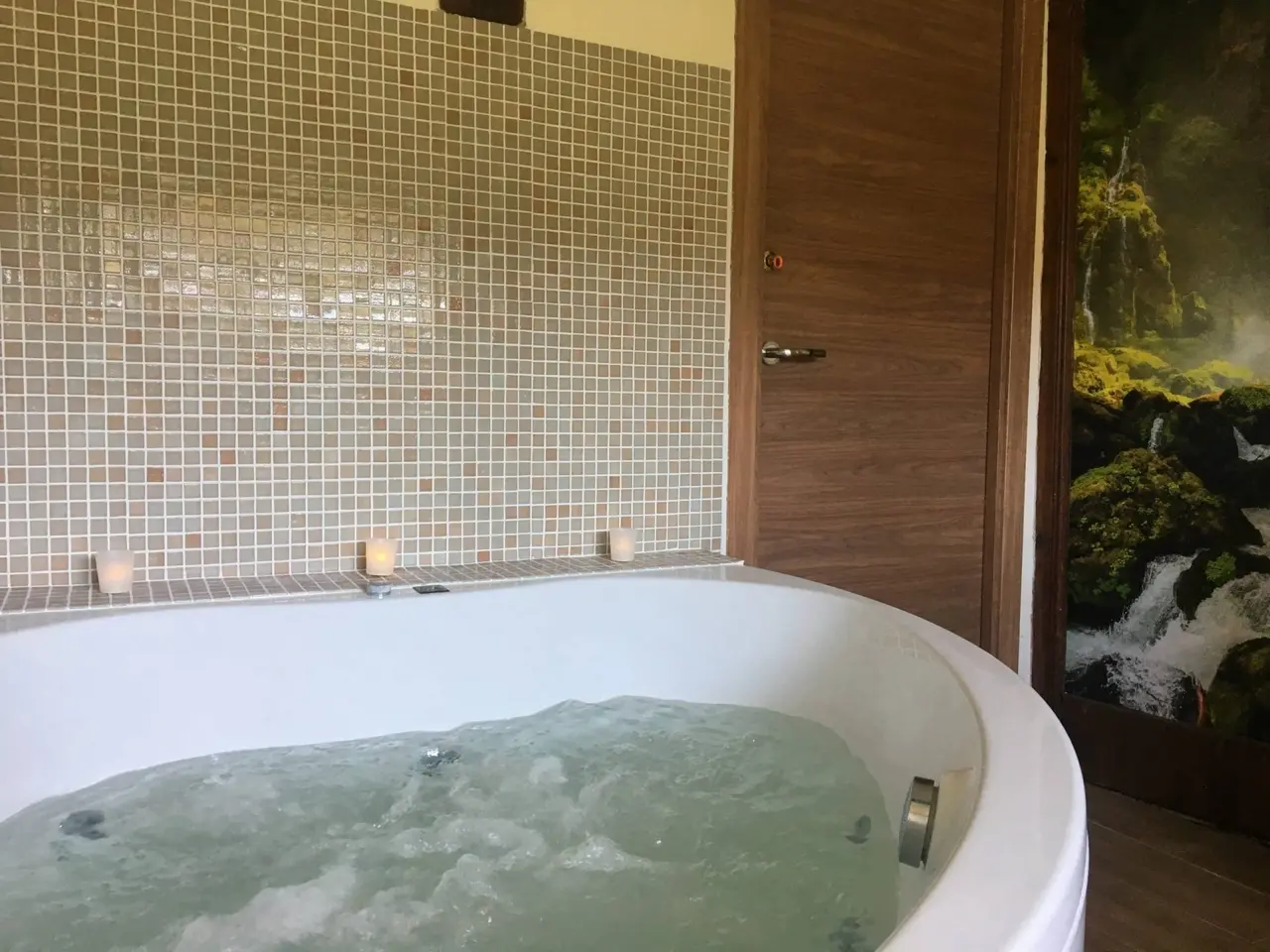 Jacuzzi privado circular de hidromasaje con agua burbujeante, rodeado de pared de mosaico beige, tres velas encendidas