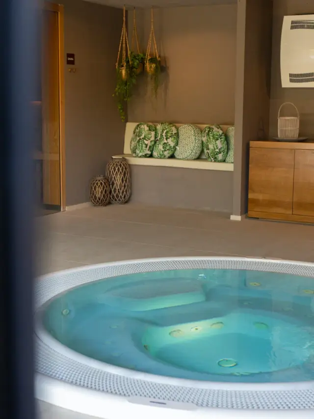 Jacuzzi circular con agua azul en un spa privado con banco acolchado, cojines verdes, plantas colgantes y muebles