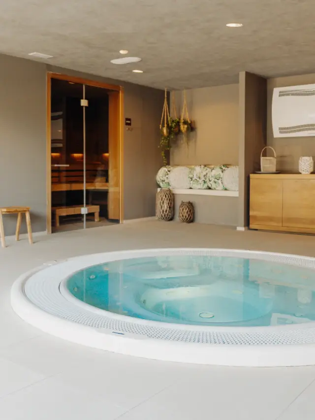 Jacuzzi circular en un spa privado con sauna visible al fondo y zona de relax con toallas y plantas colgantes.