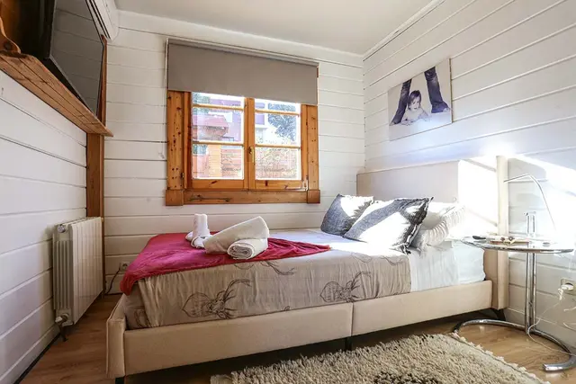 Habitación con cama doble, ventana de madera, paredes de listones blancos y muebles claros.
