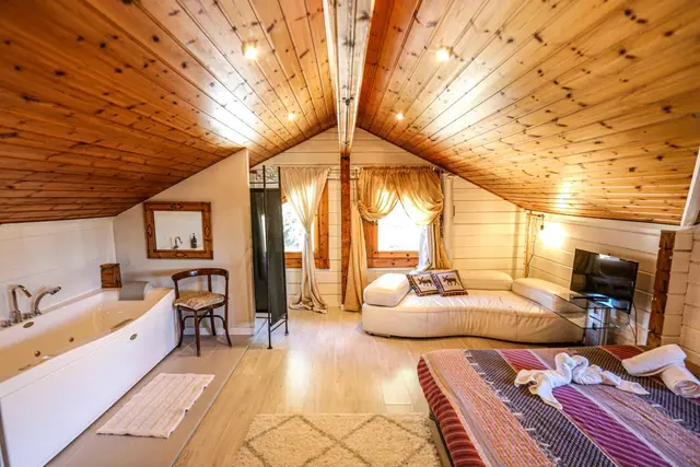 Habitación abuhardillada con techo de madera, bañera de hidromasaje blanca a la izquierda y cama grande al fondo.