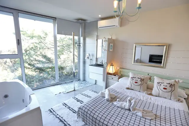 Habitación con cama doble, bañera de hidromasaje blanca en primer plano y gran ventana con vistas a la vegetación.