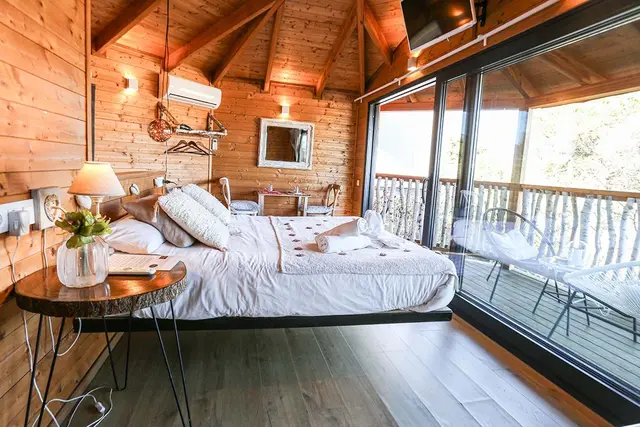Dormitorio de cabaña de madera con cama grande, mesa auxiliar y puertas de vidrio que dan a una terraza exterior.