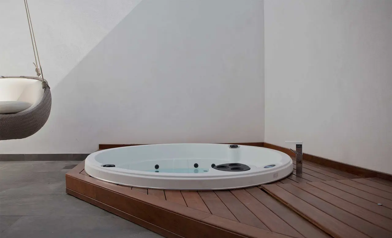 Jacuzzi circular blanco con agua, instalado sobre una plataforma de madera en un espacio exterior con paredes blancas.
