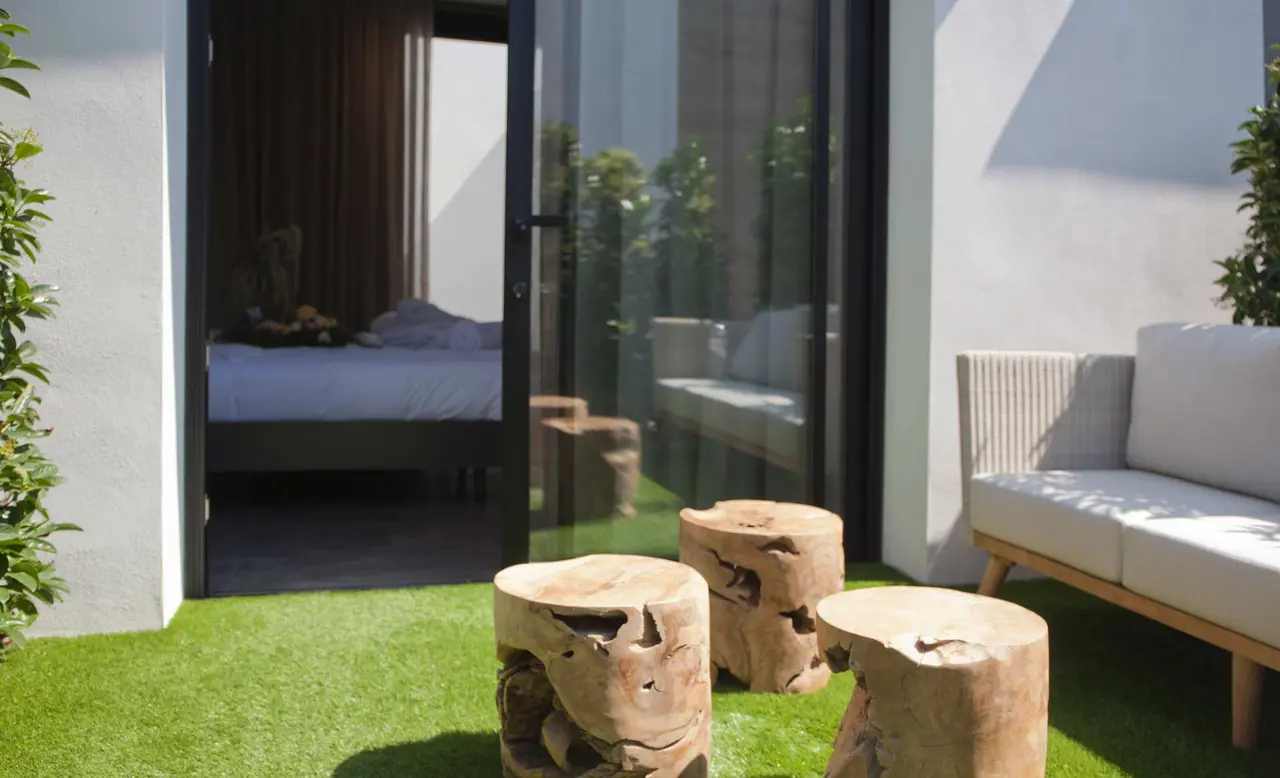 Vista exterior de una terraza con césped artificial, tres troncos de madera como mesas auxiliares, un sofá exterior