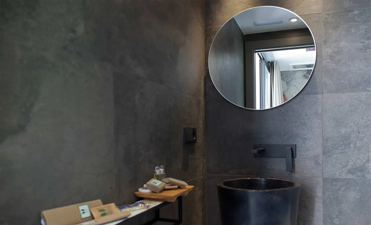 Baño con paredes de azulejos grises, lavabo negro redondo sobre pedestal, grifo negro mural, espejo circular