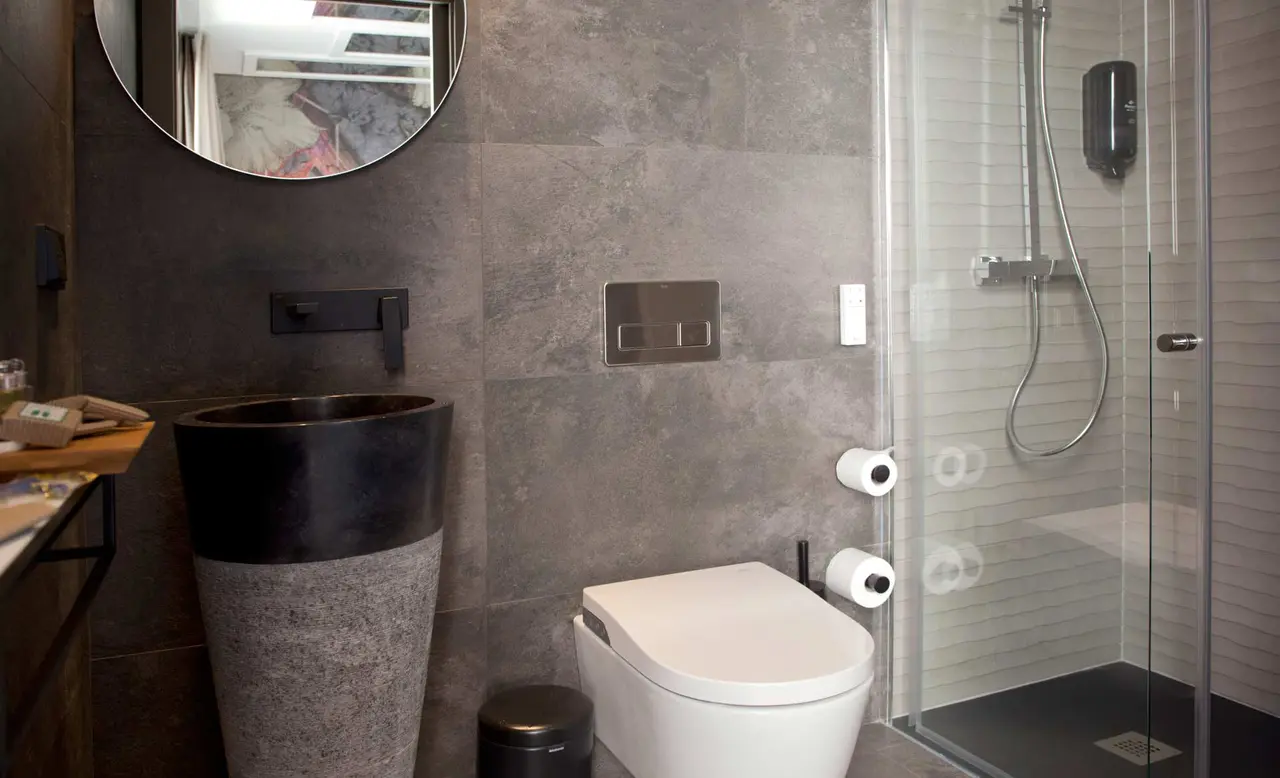 Baño con paredes de azulejos grises, inodoro blanco, lavabo de piedra alto de color negro y gris, espejo redondo