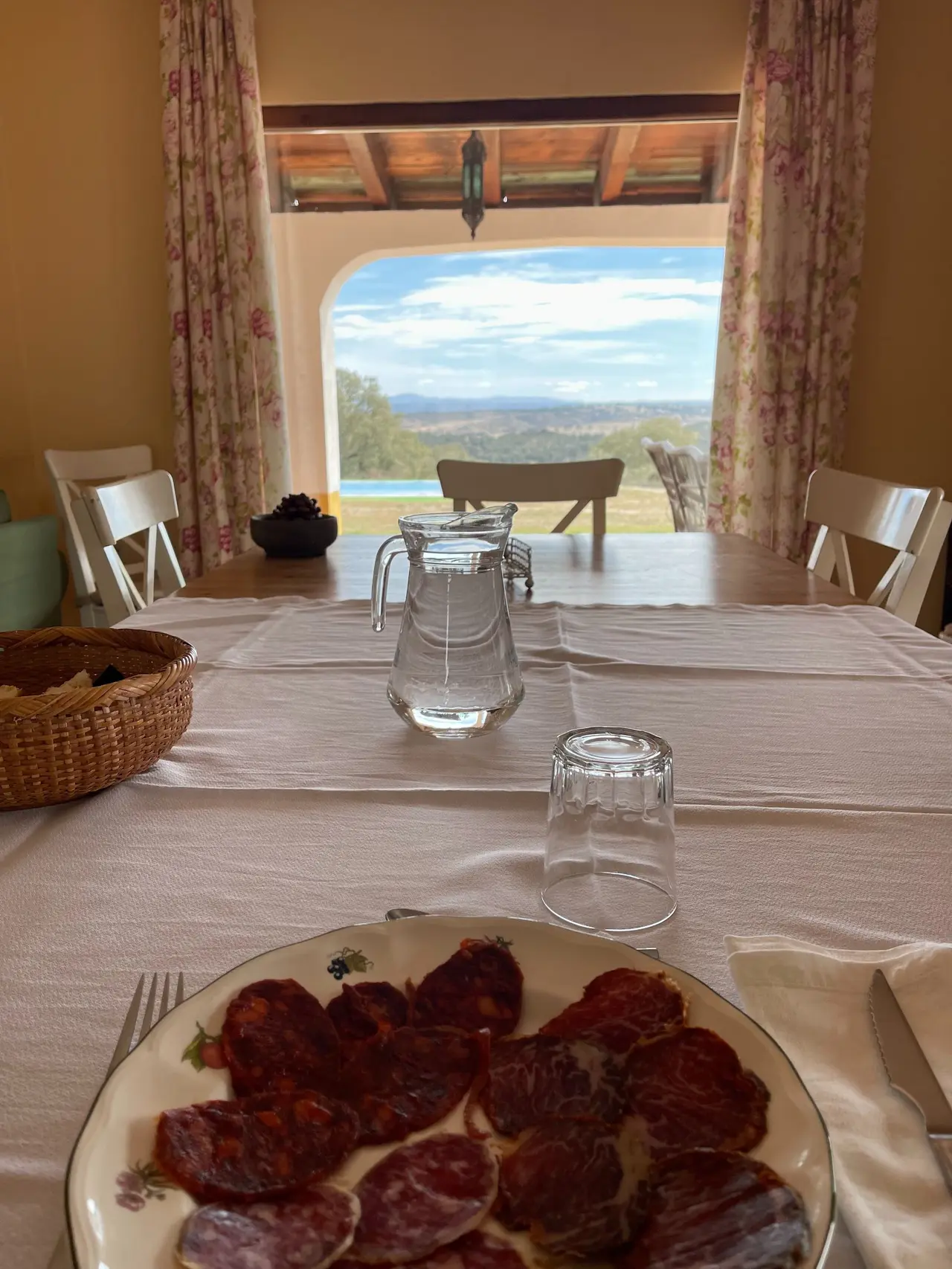 Vista desde el interior de un comedor con mesa puesta con plato de embutidos, jarra de agua y vaso, hacia una ventana