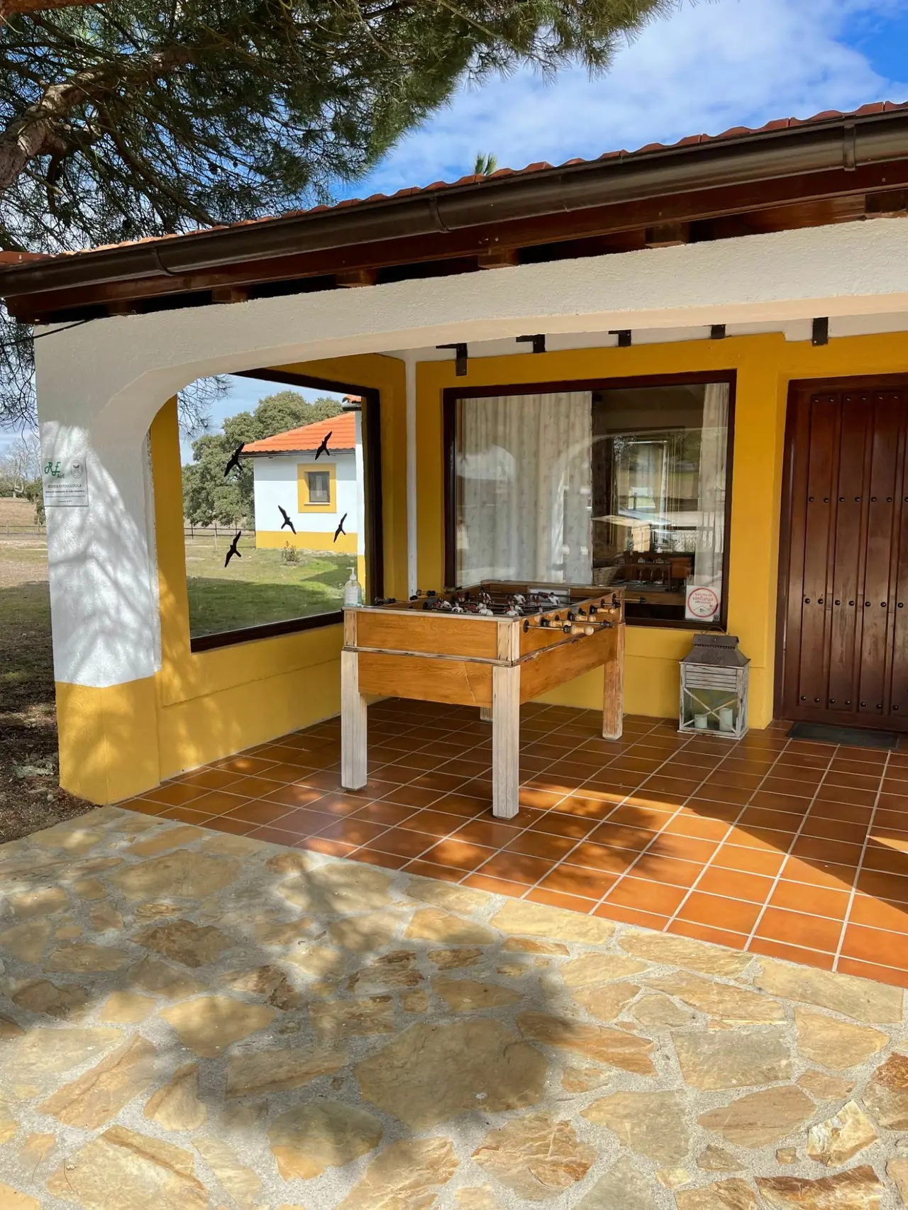 Vista exterior de una terraza cubierta con suelo de terracota y piedra, donde hay una mesa de futbolín de madera bajo