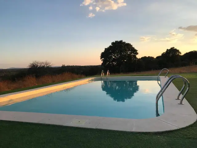 Piscina rectangular exterior con bordes de piedra clara y barandillas metálicas para acceso, rodeada de césped.