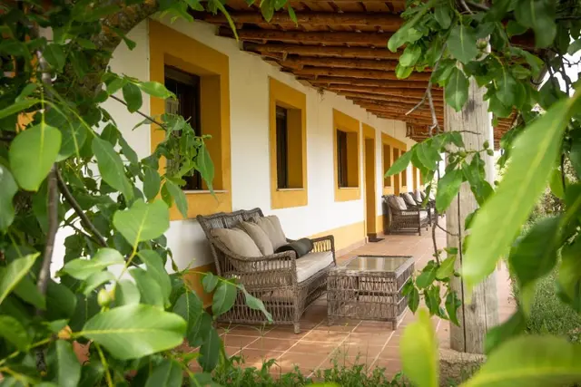 Porche exterior de una casa rural con paredes blancas y marcos amarillos, suelo de terrazo, muebles de mimbre