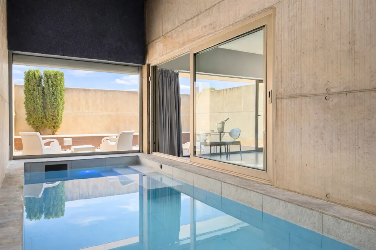 Piscina privada interior con agua azul en suite con grandes ventanales al exterior y paredes de hormigón.