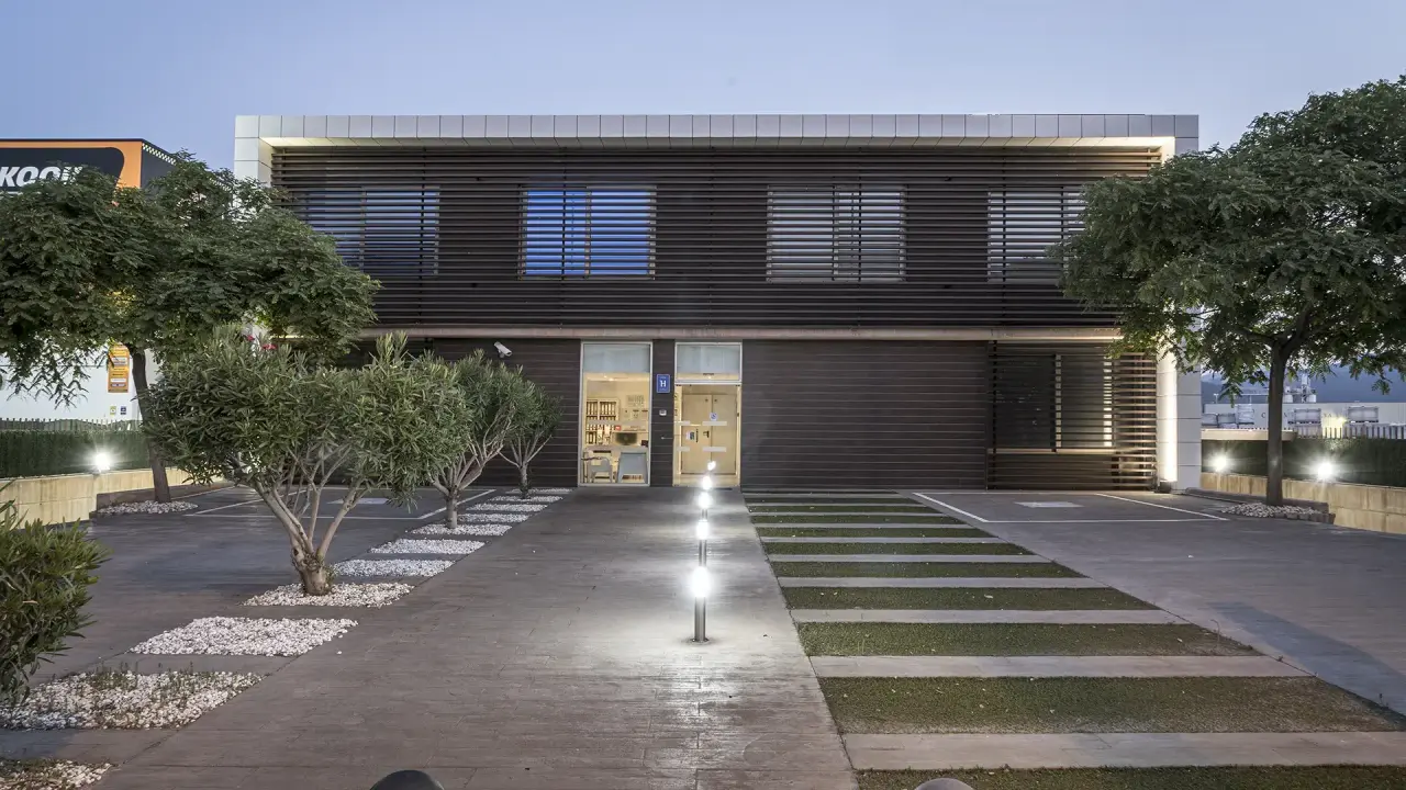Fachada exterior de un edificio moderno de dos plantas con entrada iluminada, jardín y camino de acceso al atardecer.