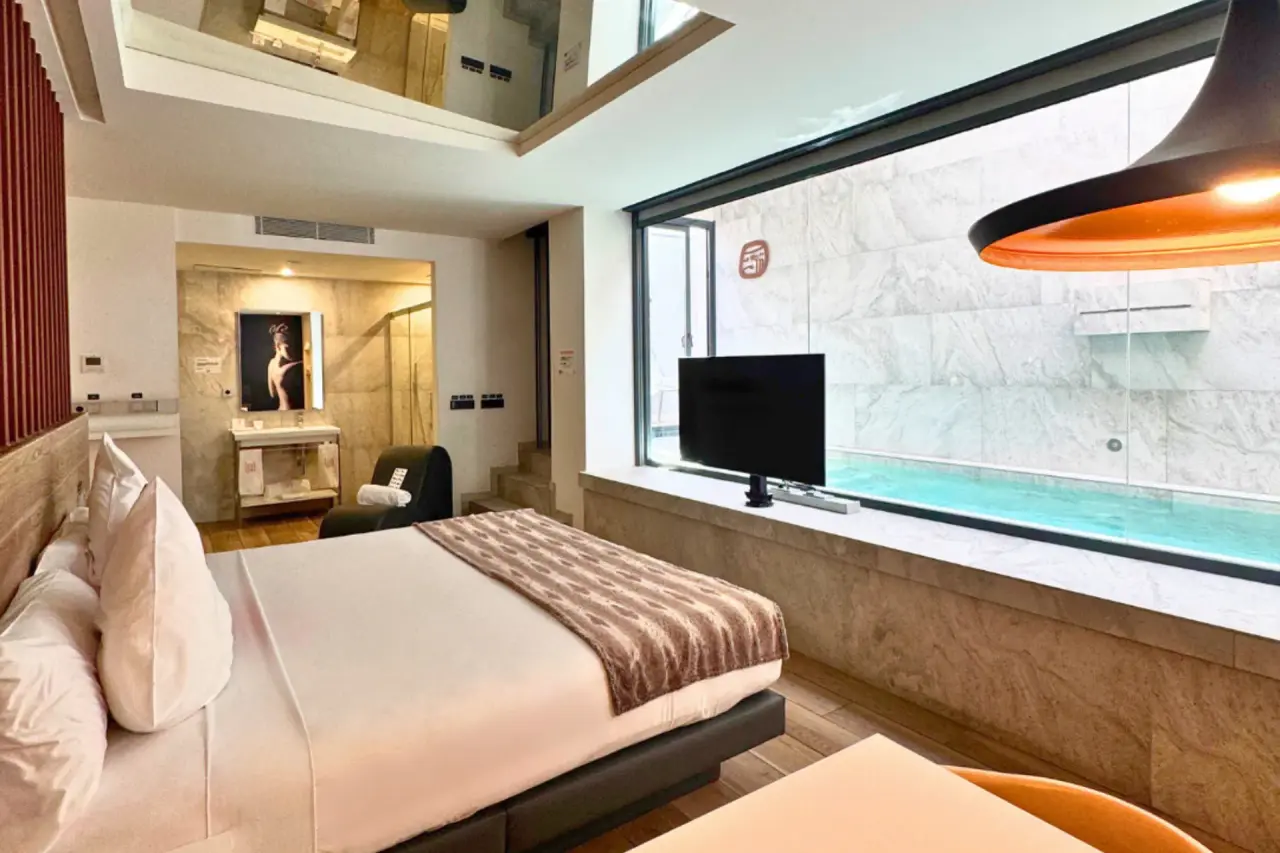 Habitación de suite con cama grande, ventana panorámica a piscina privada y zona de baño visible al fondo.