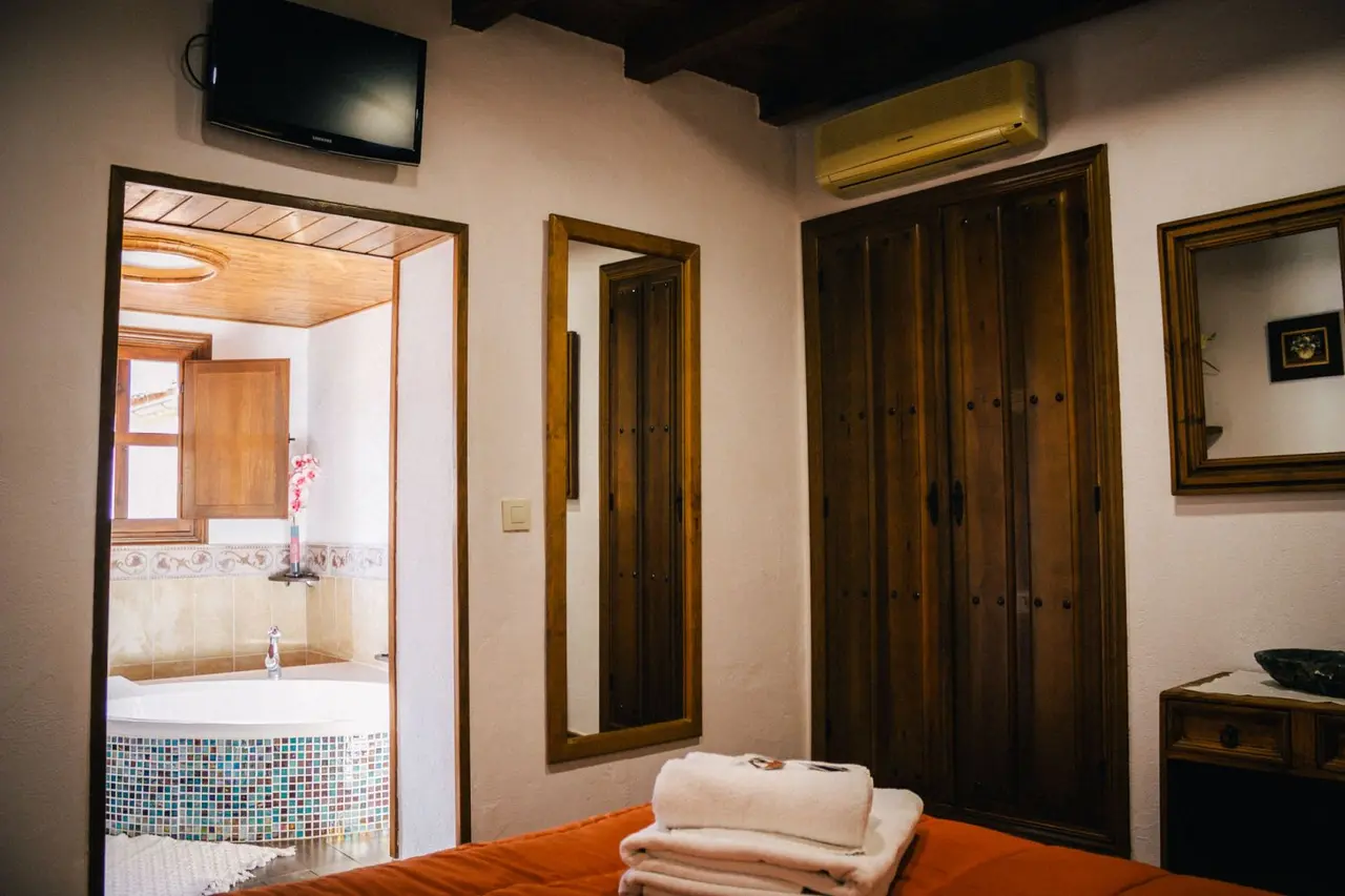 Vista de una habitación con cama en primer plano, armario de madera, espejo y televisor en la pared.