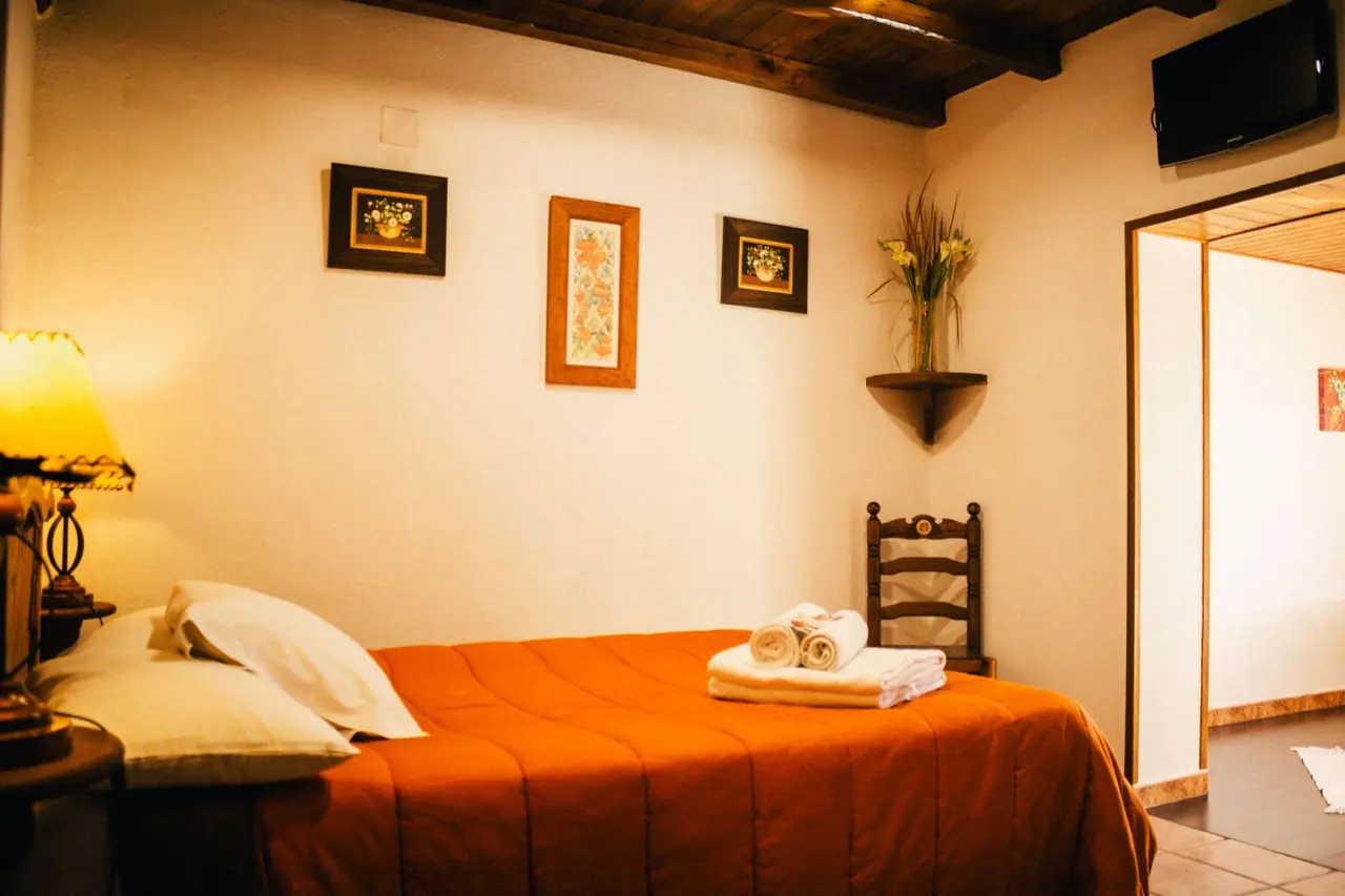 Habitación con cama doble cubierta por una colcha naranja, dos almohadas blancas, toallas enrolladas, mesita de noche