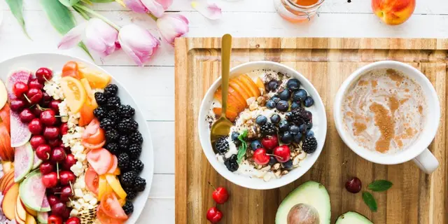 Desayuno variado con yogur, frutas, bayas y café sobre tabla de madera con flores y aguacate.