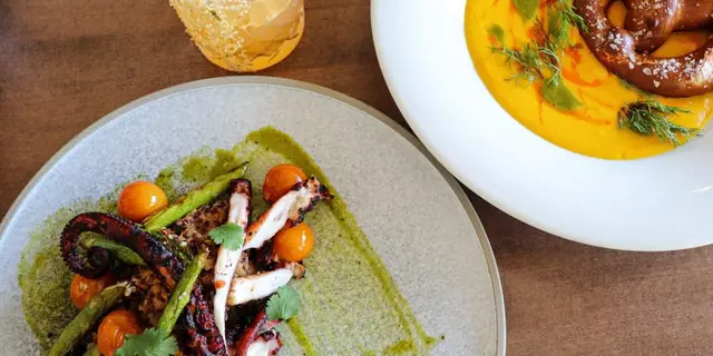 Plato de pulpo a la plancha con tomates cherry y salsa verde junto a un plato de sopa de calabaza con salchicha