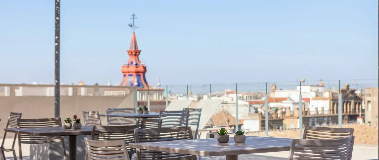 Terraza con mesas y sillas de madera frente a una vista urbana con una torre roja destacada en el horizonte.