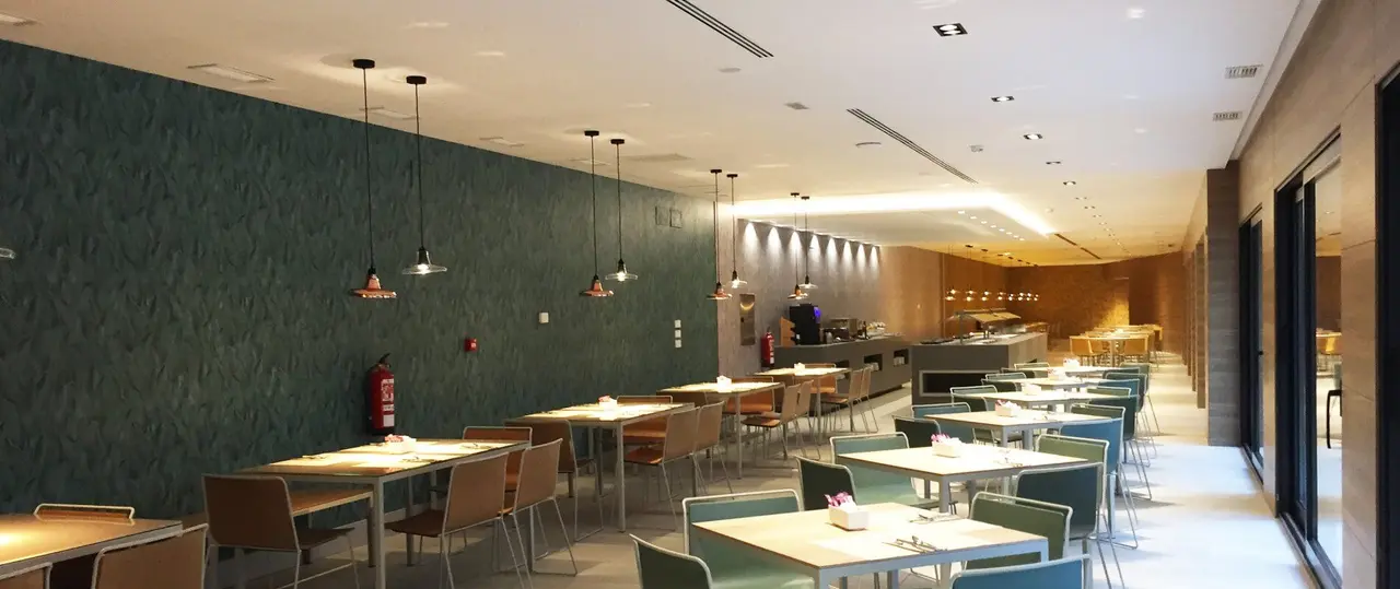 Interior de un comedor de hotel con mesas y sillas dispuestas en filas, paredes verdes y zona de buffet al fondo.
