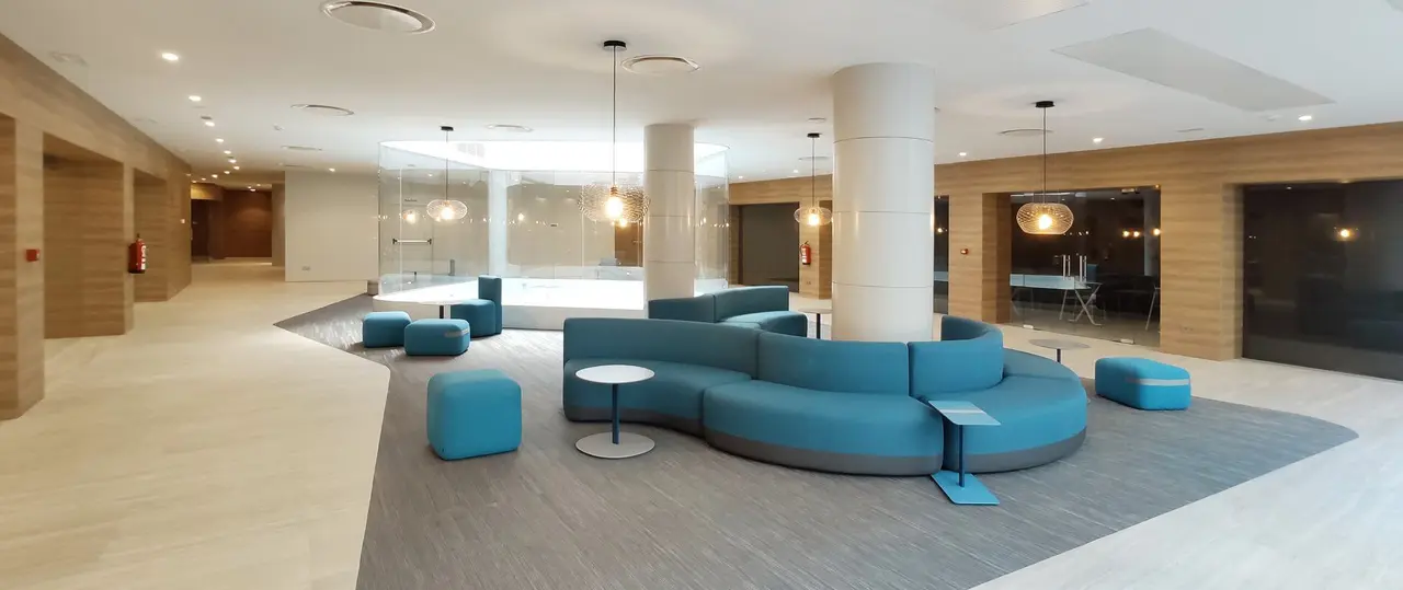 Amplio lobby de hotel con sofás curvos azules, mesas auxiliares y pilares blancos en un espacio moderno y luminoso.