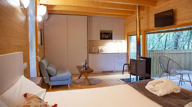 Interior de una cabaña de madera con cama grande en primer plano, sofá azul, mesa de centro, cocina equipada al fondo