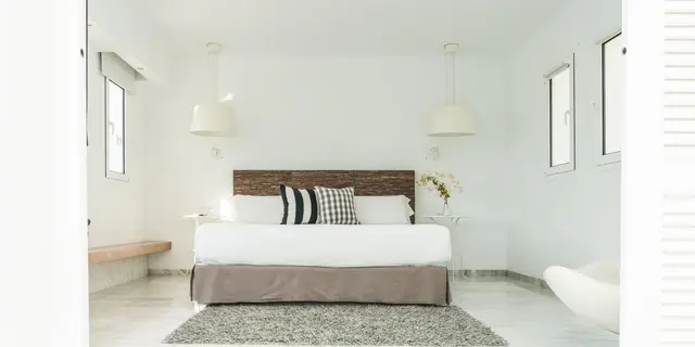 Vista de una habitación luminosa y minimalista con una cama grande vestida de blanco y gris, cabecero de madera