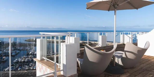Terraza exterior con suelo de madera, dos sillones modernos y una sombrilla, con barandilla de vidrio que ofrece
