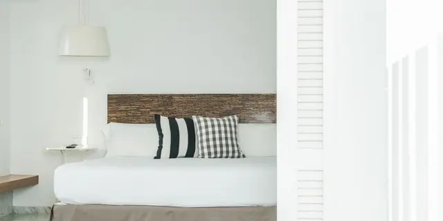 Vista parcial de una habitación con cama blanca, cabecero de madera, almohadas decorativas y lámpara colgante, vista