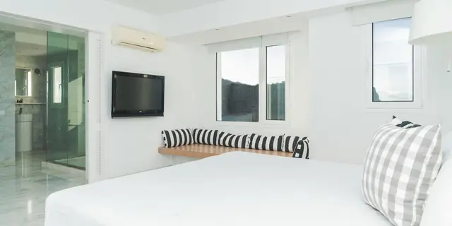 Habitación blanca con cama grande, ventana con banco acolchado a rayas, televisor en la pared, aire acondicionado