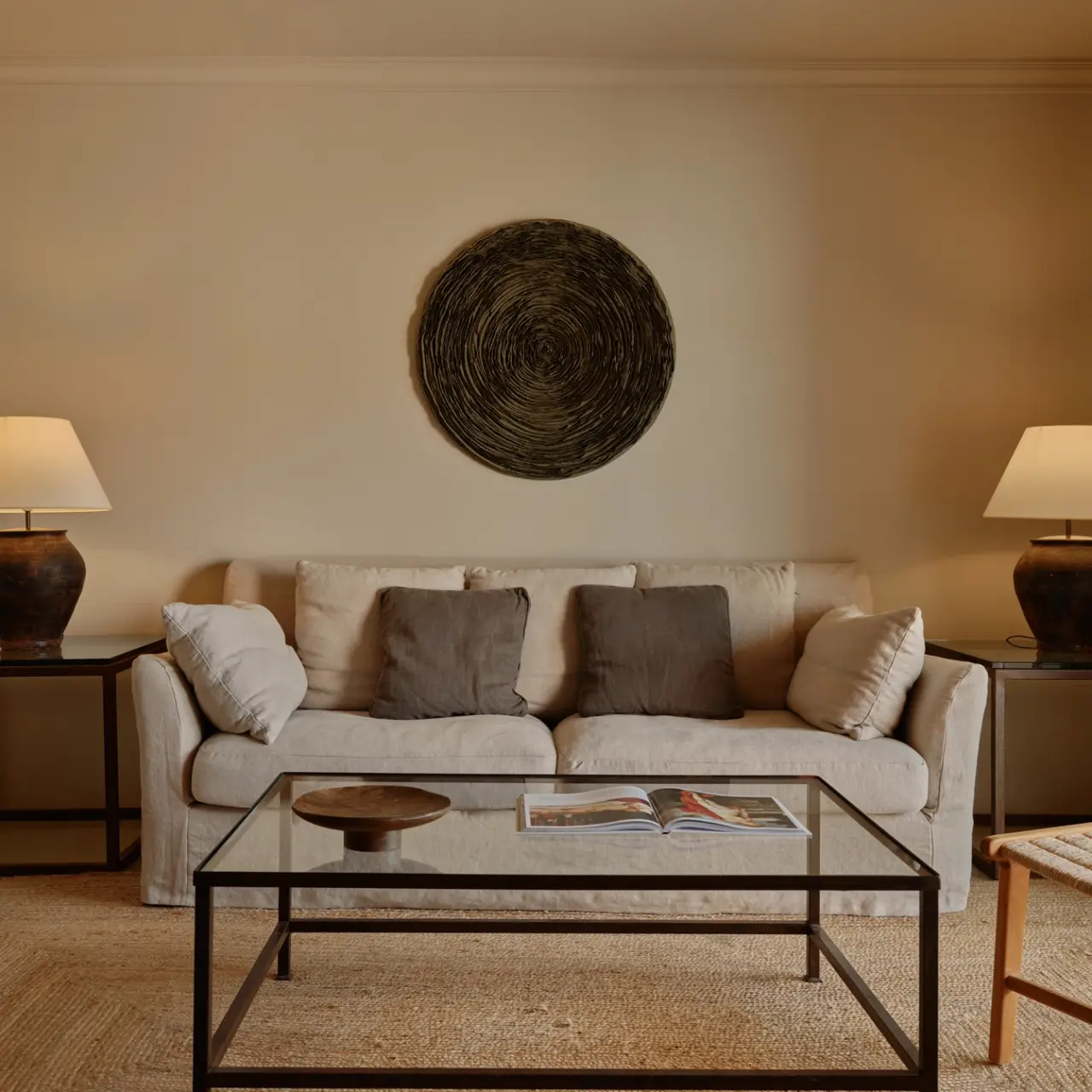 Sala de estar con sofá beige de tres plazas, cojines grises y blancos, mesa de centro de cristal y metal, dos lámparas