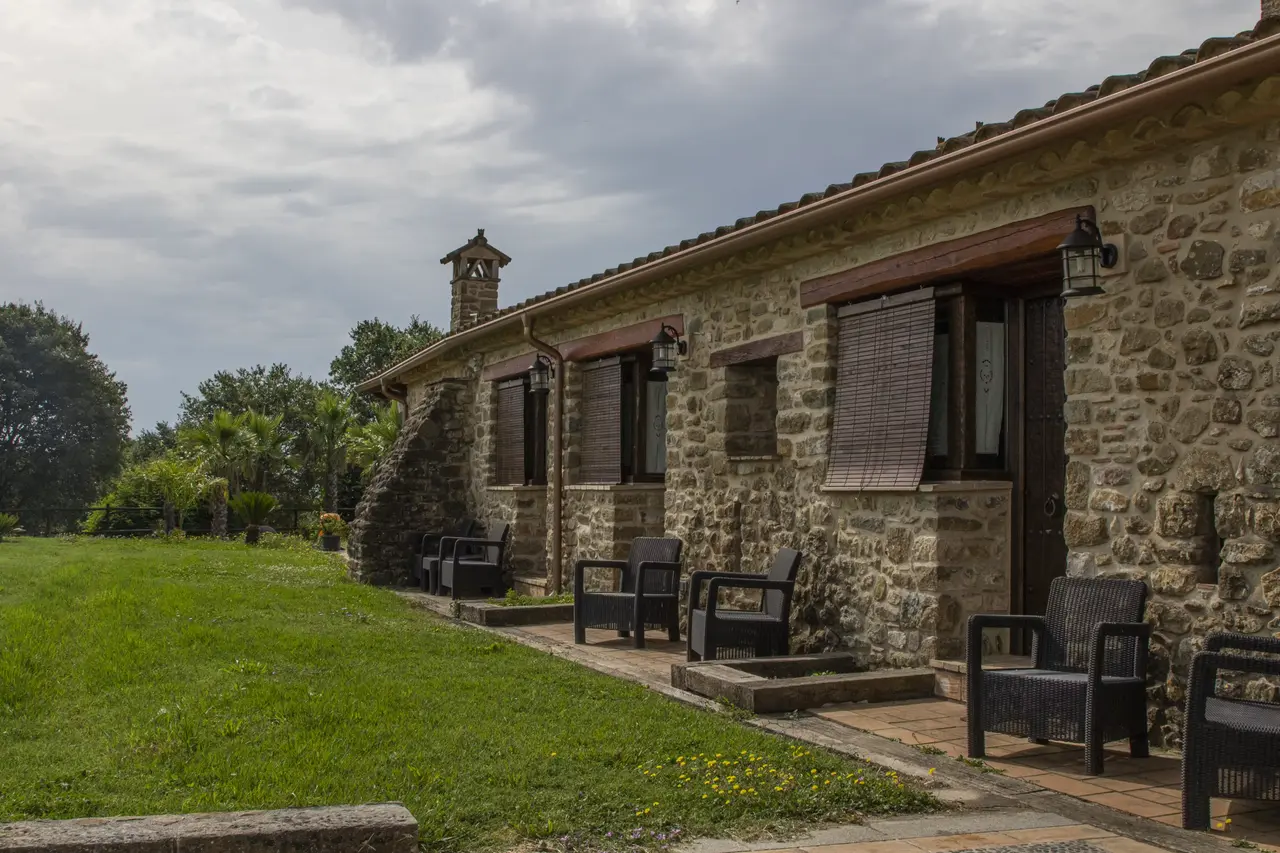 Fachada lateral de una casa rural de piedra con varias puertas y ventanas, terraza con sillas de exterior y un amplio
