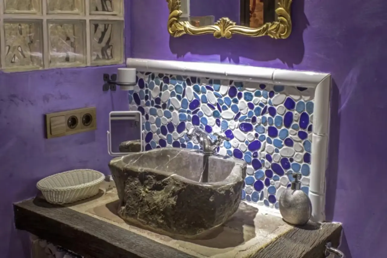 Baño con paredes moradas, lavabo de piedra irregular sobre encimera de madera, grifo plateado, mosaico de piedras