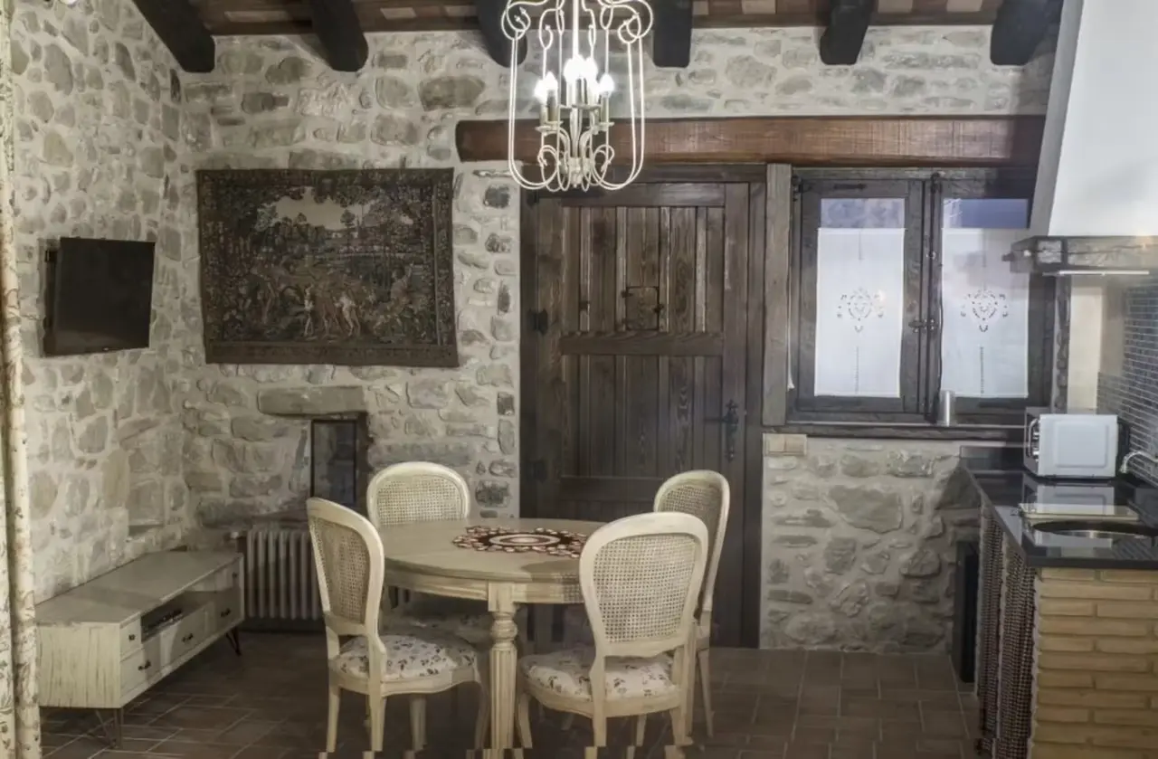 Interior de una habitación con paredes de piedra vista, mesa redonda de comedor con cuatro sillas tapizadas, televisor