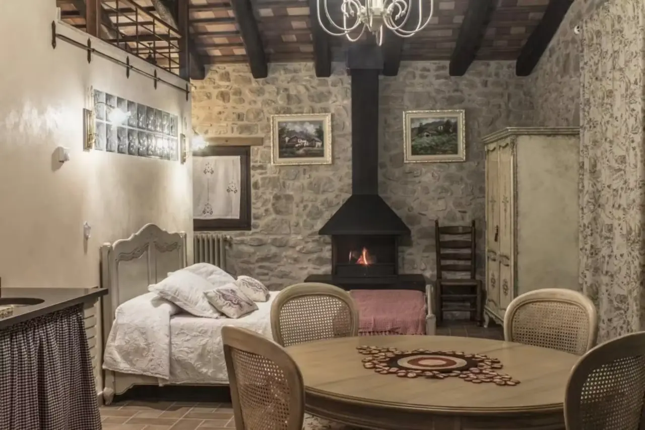 Interior de una habitación rústica con paredes de piedra, techo de vigas de madera, cama con cabecero blanco y ropa