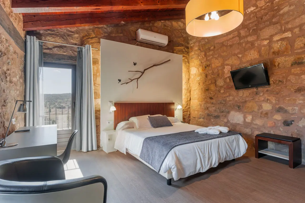 Habitación suite con cama doble, paredes de piedra vista, vigas de madera en el techo, escritorio, silla, televisor