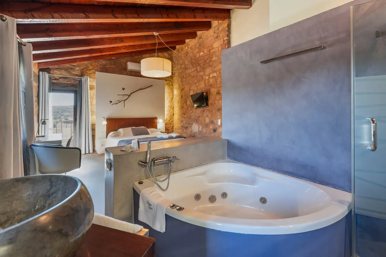 Jacuzzi blanco de hidromasaje en primer plano dentro de una suite rústica con paredes de piedra y vigas de madera.
