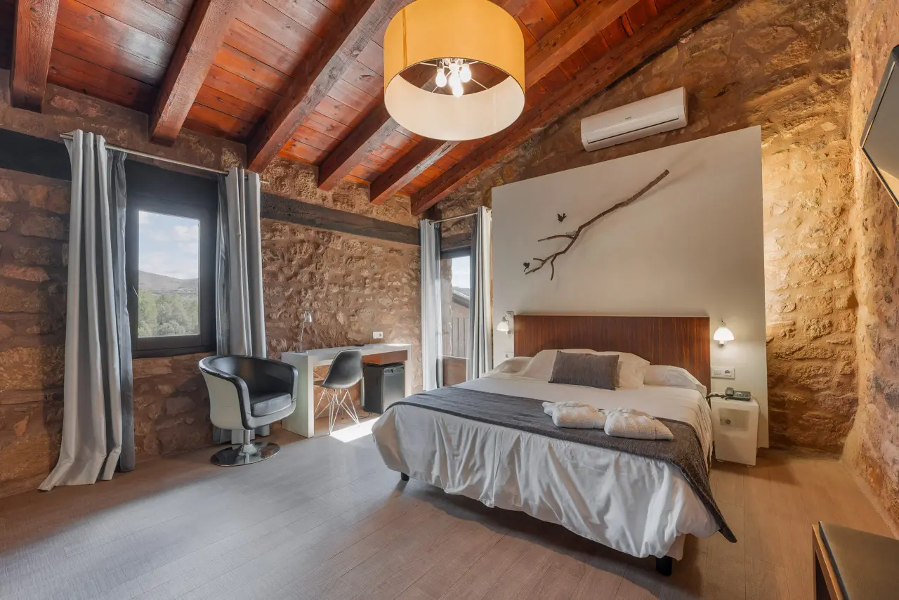 Habitación suite con cama doble, cabecero de madera, paredes de piedra vista, techo con vigas de madera, escritorio