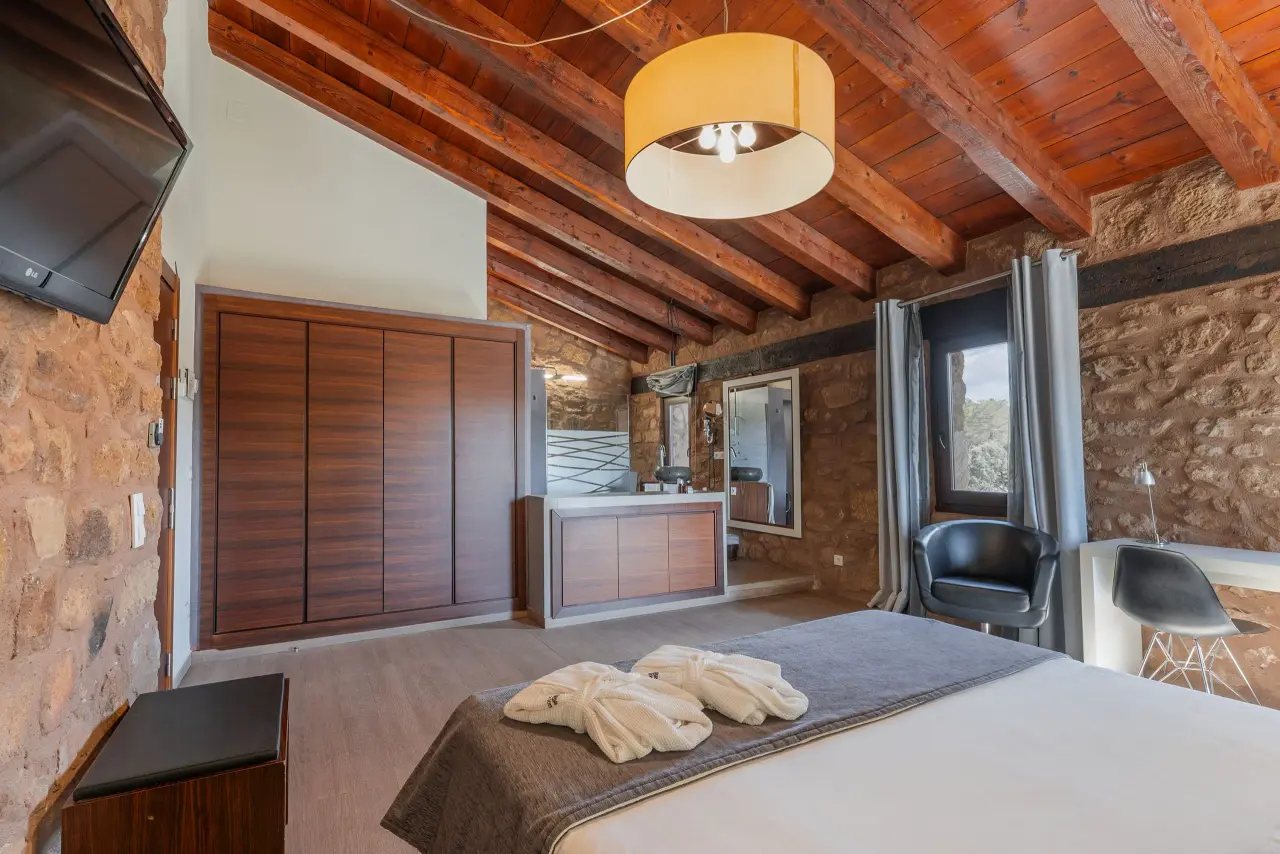 Vista de una suite con cama grande en primer plano, armario de madera, paredes de piedra y techo con vigas de madera.