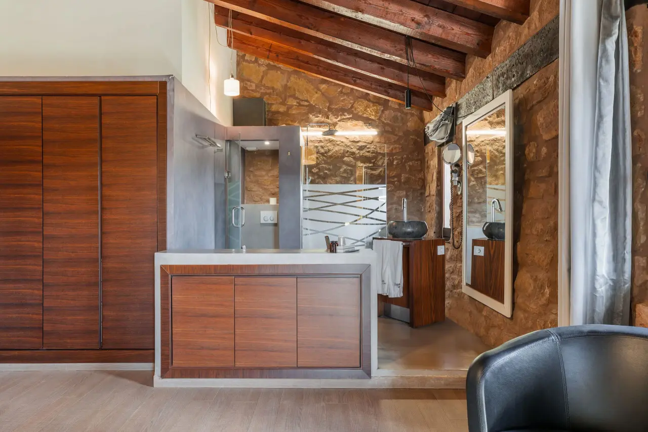Interior de un baño con paredes de piedra vista, techo con vigas de madera, mueble de lavabo doble de madera, espejo