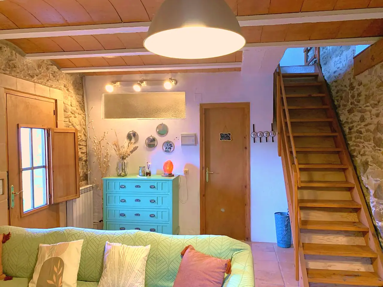 Interior de una sala con sofá verde en primer plano, cómoda azul turquesa, escalera de madera a la derecha, puerta