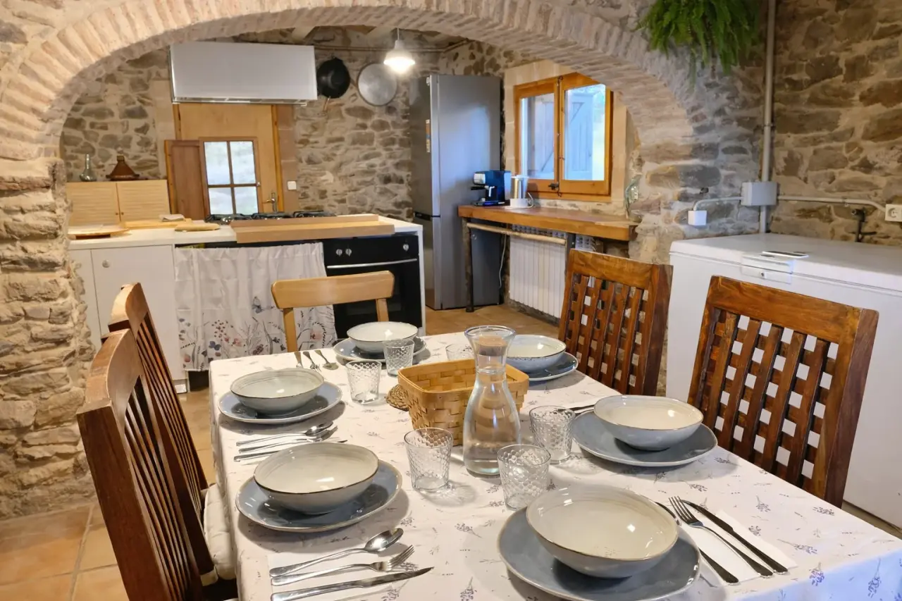Vista de un comedor rústico con mesa grande puesta para seis personas, sillas de madera y mantel blanco con estampado.