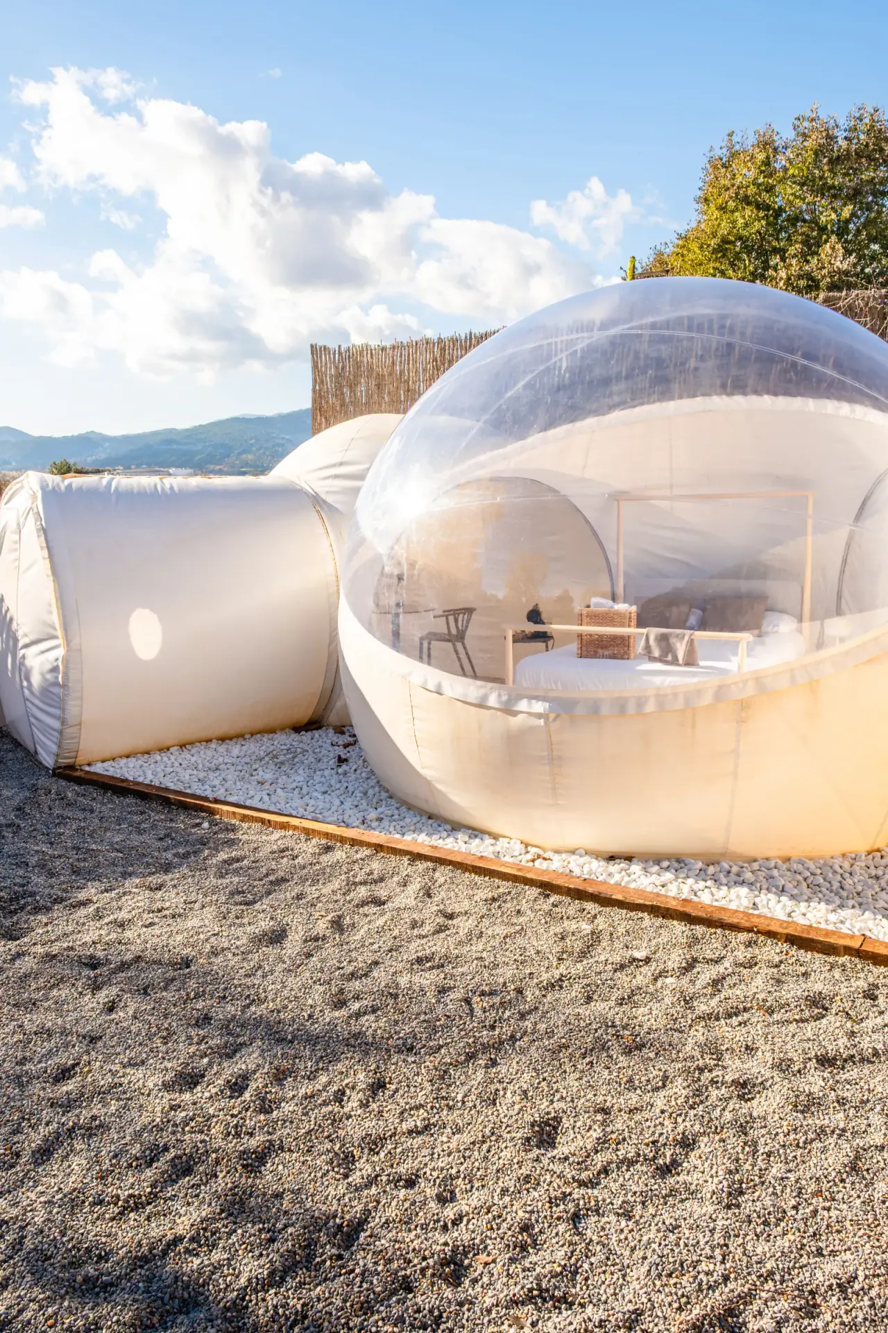 Vista exterior de una cúpula inflable transparente tipo burbuja con cama interior, situada sobre grava en un entorno