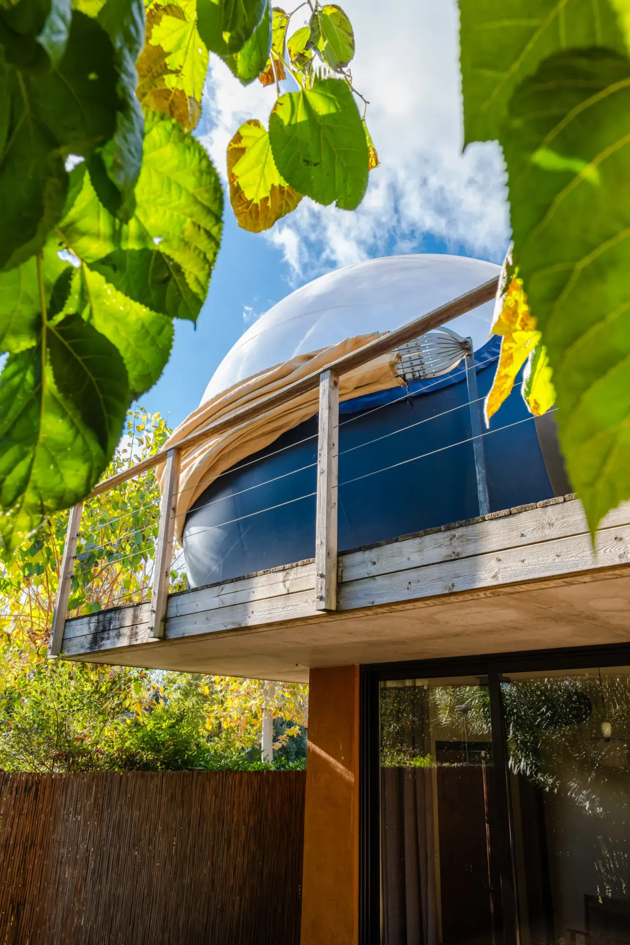 Vista exterior de una cúpula blanca elevada sobre una estructura de madera con barandilla, rodeada de follaje verde