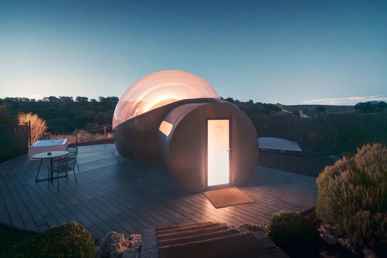 Vista exterior nocturna de una habitación tipo burbuja con cúpula transparente iluminada, situada sobre una terraza