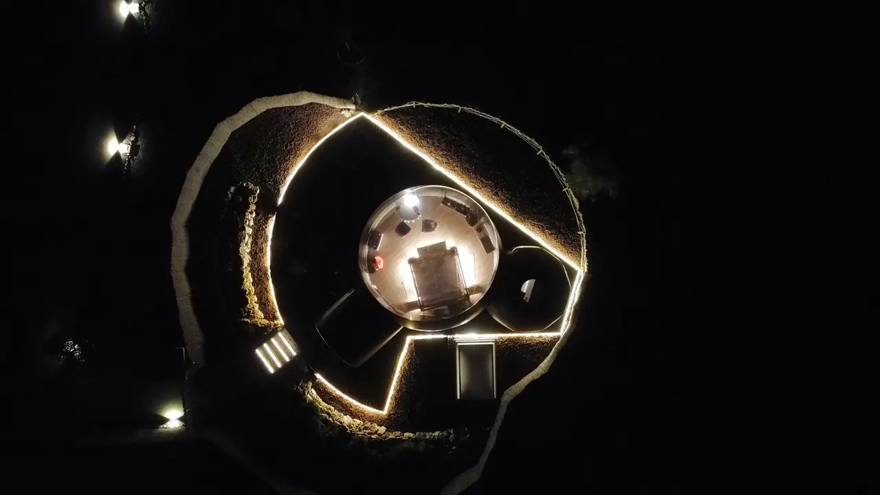 Vista aérea nocturna de una estructura circular iluminada con una esfera de vidrio en el centro, rodeada de vegetación