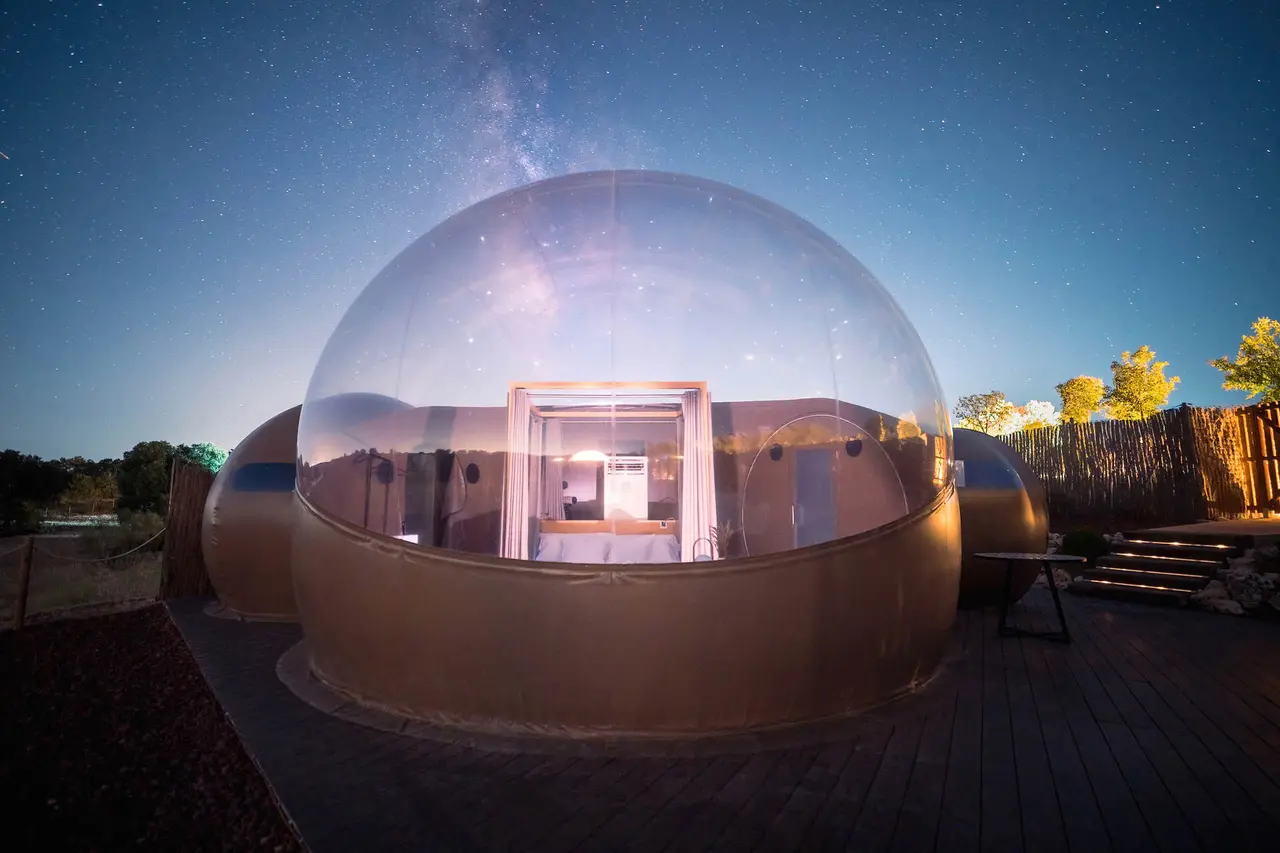 Vista nocturna de una habitación burbuja con cúpula transparente en un entorno natural, con el cielo estrellado