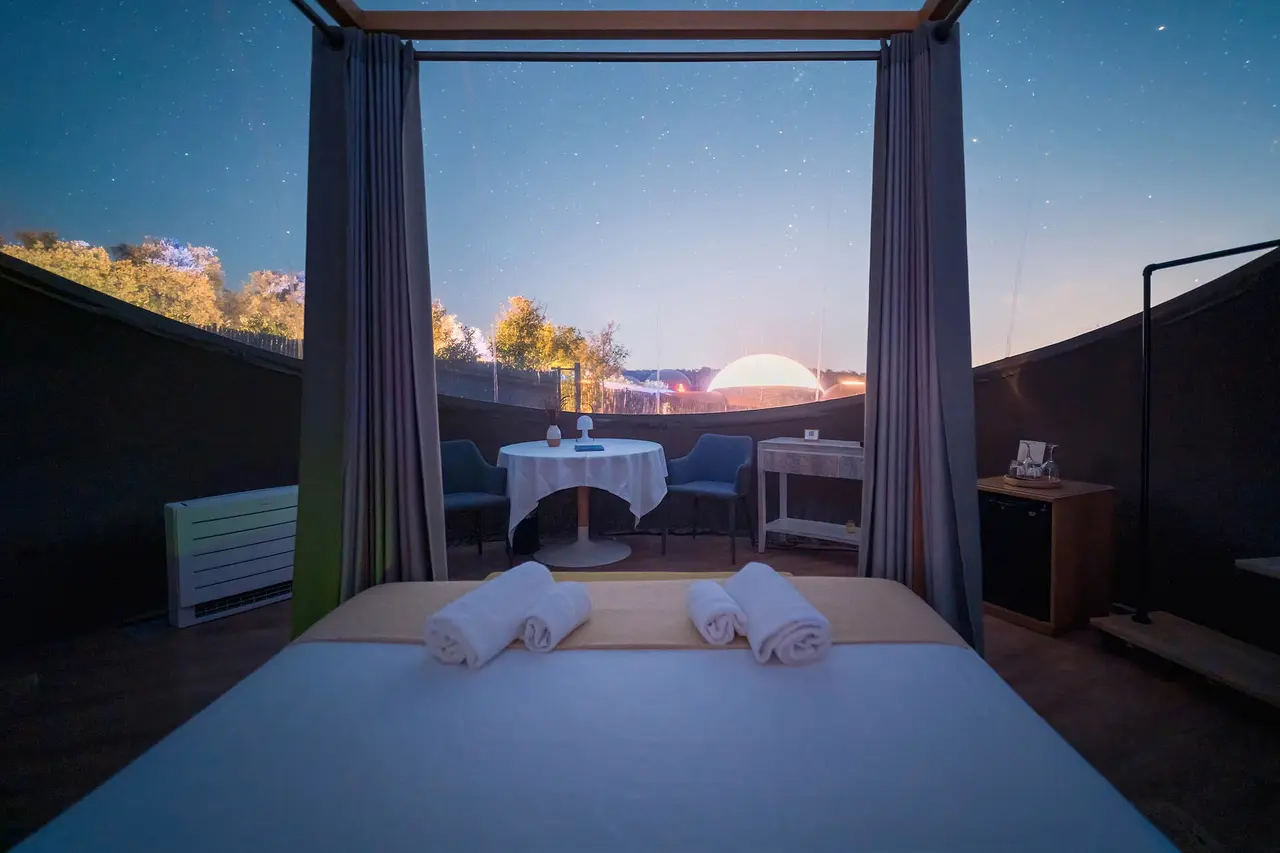 Vista interior de una habitación con cama grande en primer plano, toallas enrolladas y cortinas abiertas