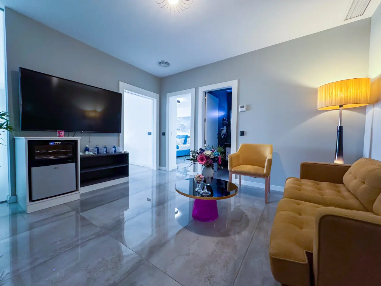 Vista de una suite de hotel con sala de estar amueblada con sofá y sillón amarillo, mesa de centro redonda, televisor