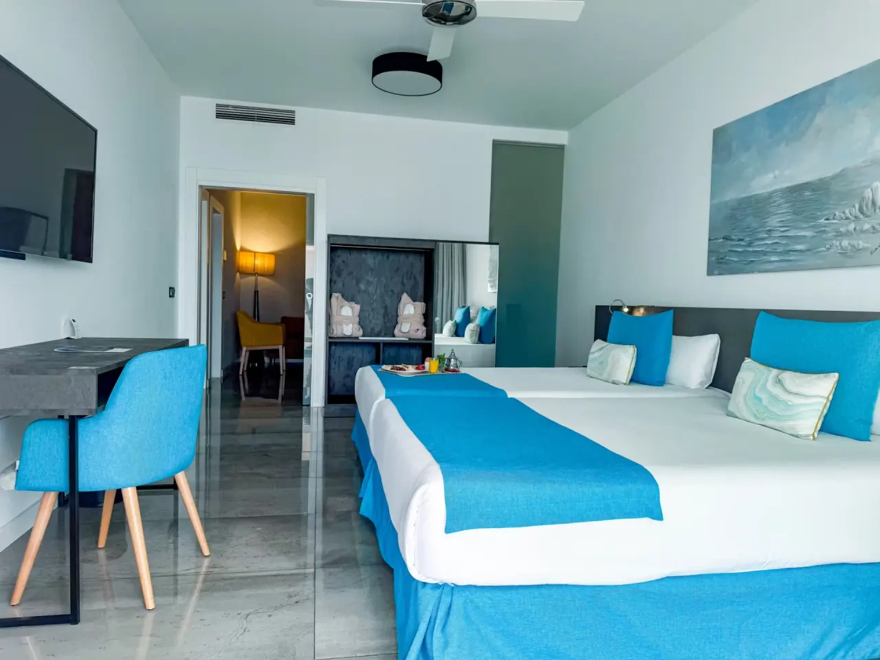 Habitación de hotel con dos camas grandes vestidas en blanco y azul, cabecero gris, almohadas decorativas, escritorio